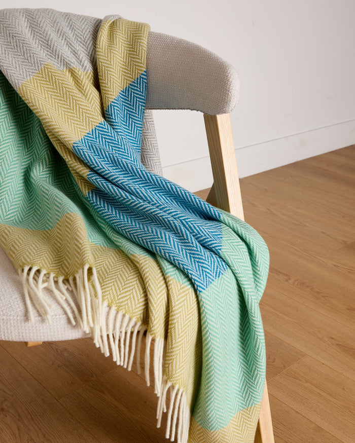 Luxurious Foxford Garavogue Cashmere and Lambswool Throw draped over a modern wooden chair. Featuring a vibrant herringbone pattern in blues, greens, and yellows, with soft fringed edges. Woven in Foxford’s historic mill in County Mayo, Ireland.